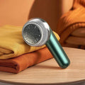 green electric lint remover placed on folded mustard and rust-colored sweaters on wooden table